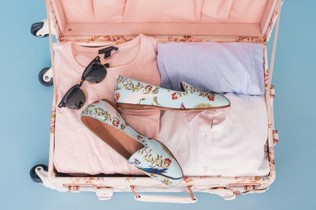 10 Nontraditional Wedding Gift Ideas for a Modern Couple, a pink suitcase filled with clothes, a pair of sunglasses, and a pair of shoes.