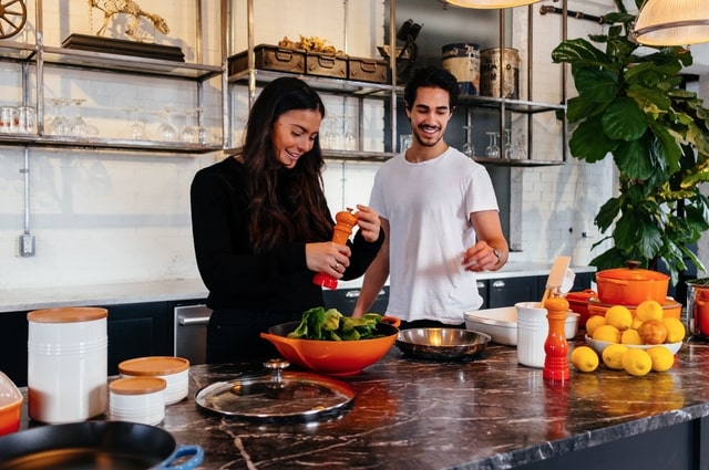 10 Nontraditional Wedding Gift Ideas for a Modern Couple, a couple in the kitchen cooking. The woman is making a salad and the man is seasoning something in a pan.