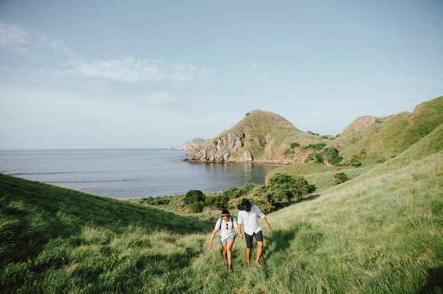 9 Tips to Make Your Wedding More Eco-Friendly, a couple hiking across a huge, grassy hill bordered by a large, beautiful body of water