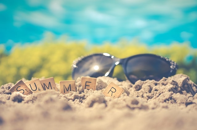 Image of a pair of sunglasses on the sand, behind scrabble tiles spelling out the word summer